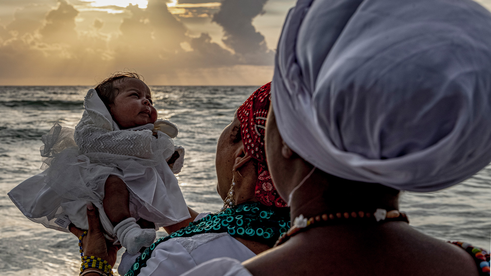 Mothers holding a child near the ocean in a religious ceremony.