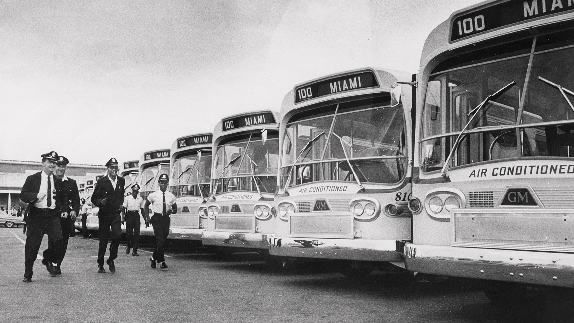 A black and white image of a group of people dressed as bus drivers run in front of a row of parked buses.