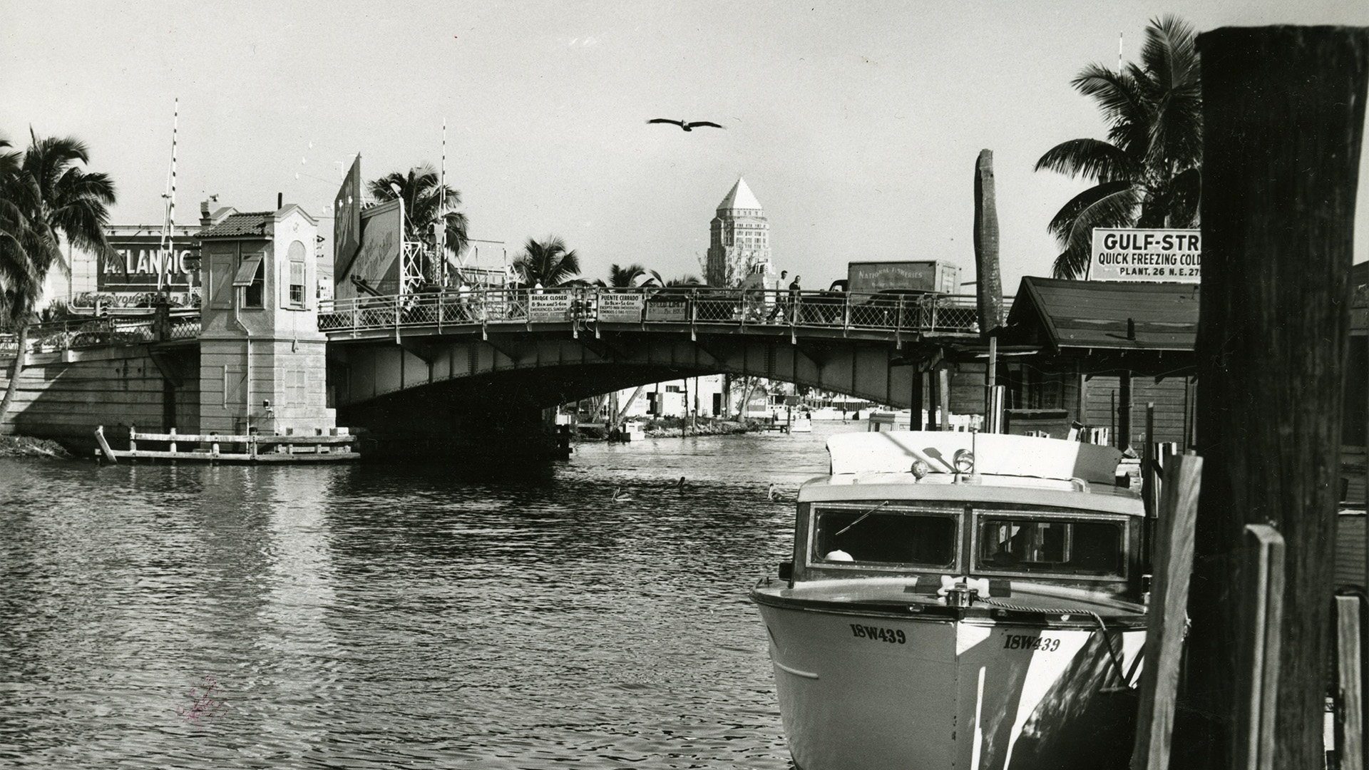 Black and white historical image of the Miami River. A boat is docked alongside the river with a bridge just behind it. In the background, a city skyline can be seen.