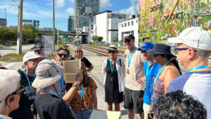 A group stands on the side of a paved road in the Design District. They look towards a tour guide who stands wearing a hat, black shirt, and shorts and is holding a piece of paper up to the group.