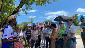 A tour guide wearing a straw hat and blue and white Haitian shirt stands in front of a group of tour participants in Little Haiti on a sunny day.