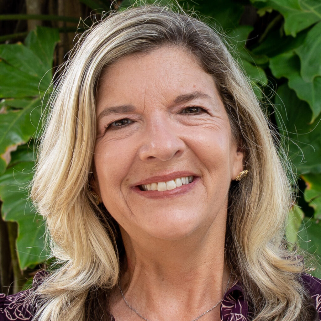 A person with wavy blonde shoulder length hair smiles at the camera. They wear gold earrings and a purple top.