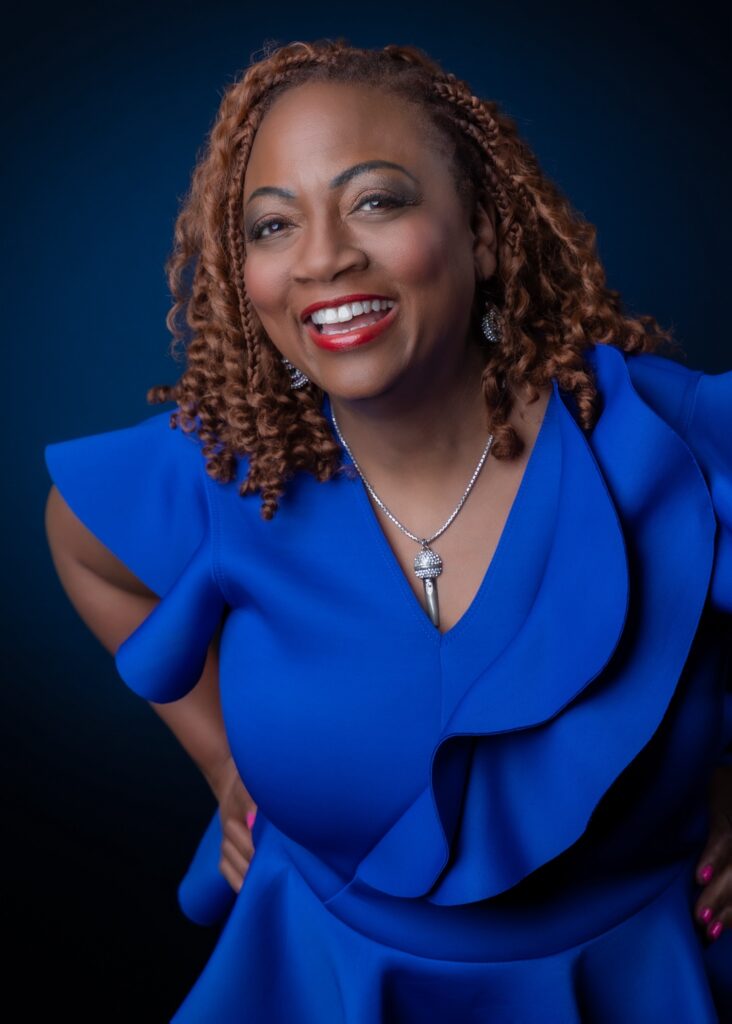 A person wearing a bright blue ruffled top, diamond earrings and necklace with a microphone motif, and red lipstick smiles at the camera with their hands on their hips.