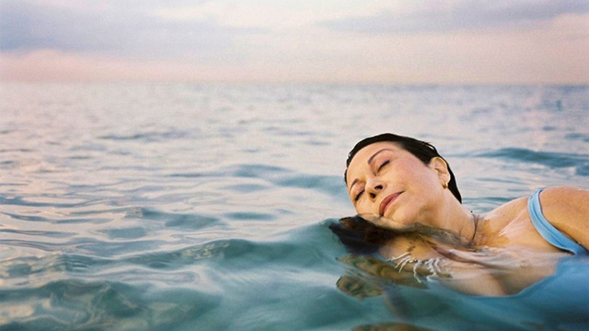 View of a person swimming in rippling open water with pastel skies behind them.