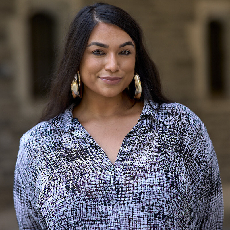 A person with long straight dark brown hair wears large gold hoop earrings and a small smile. They wear a black and white patterned blouse.