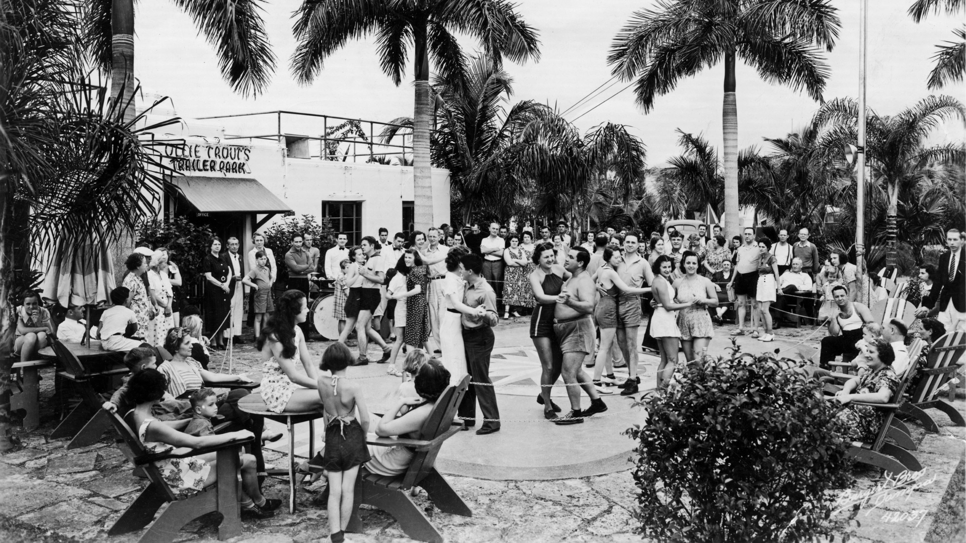 A sepia image of people dancing on a dance floor as some look on from patio chairs outdoors in front of a building with the sign 