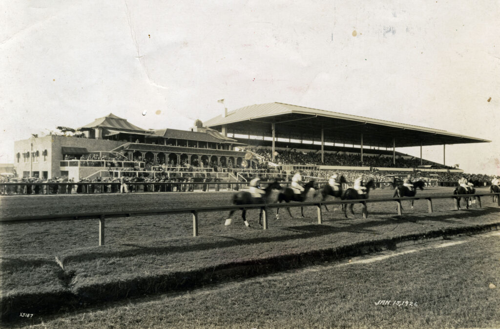 A black and white image of the Hialeah Park racetrack with spectators looking on as horses run.