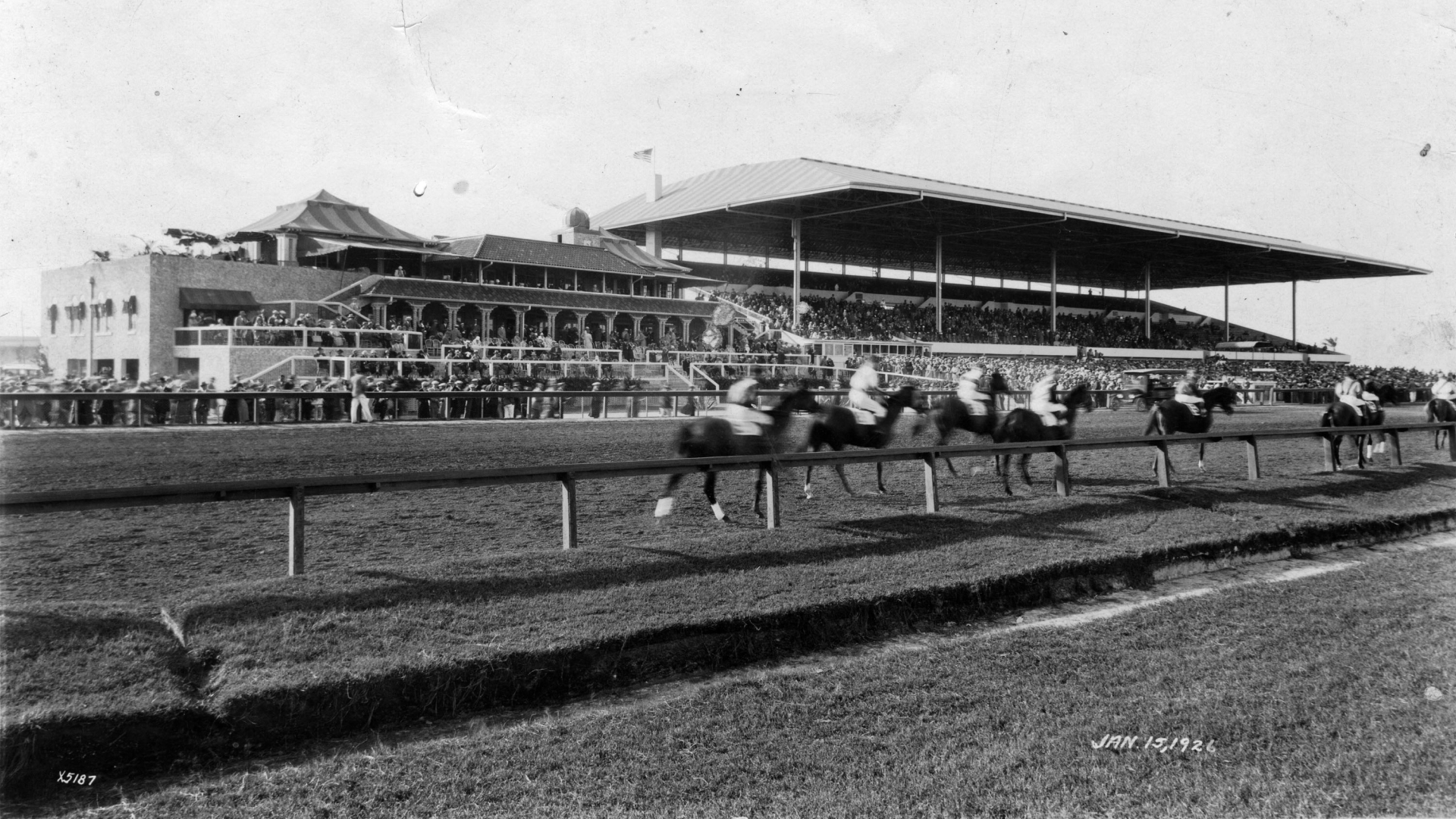 A black and white archival image of the Hialeah Park racetrack with spectators looking on as horses race.