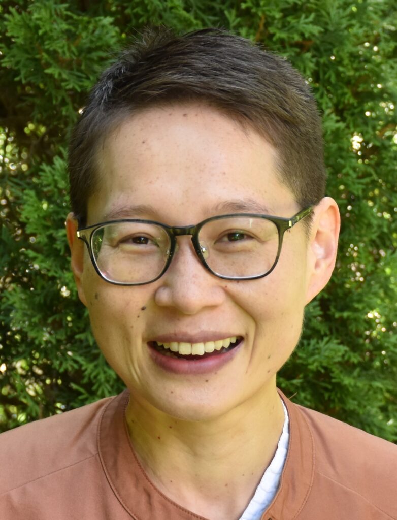 A person with short brown hair and black rimmed glasses smiles at the camera. They are wearing a muted pink top and are standing in front of green foliage.