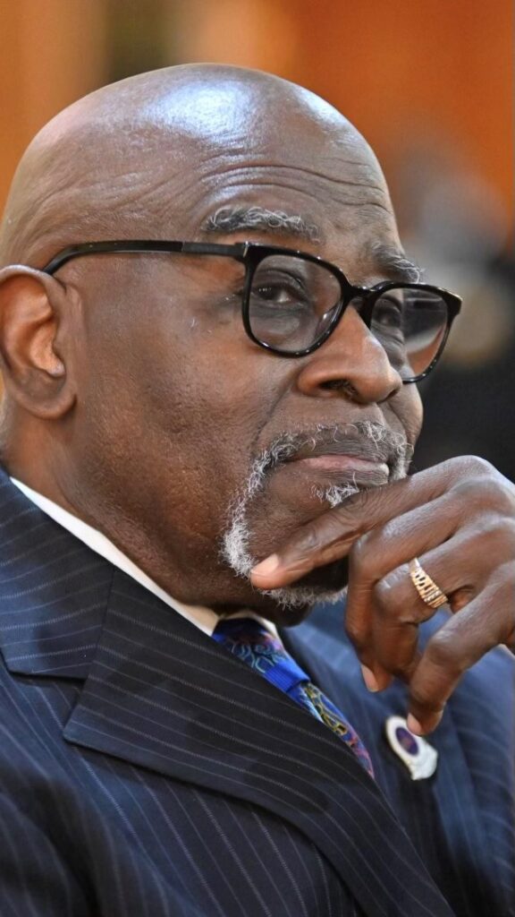 A person with black rimmed glasses and a gold ring smiles faintly as they gaze away from the camera. They have their hand on their chin and wear a gold ring, pinstriped navy suit, and bright blue tie with yellow and red accents.