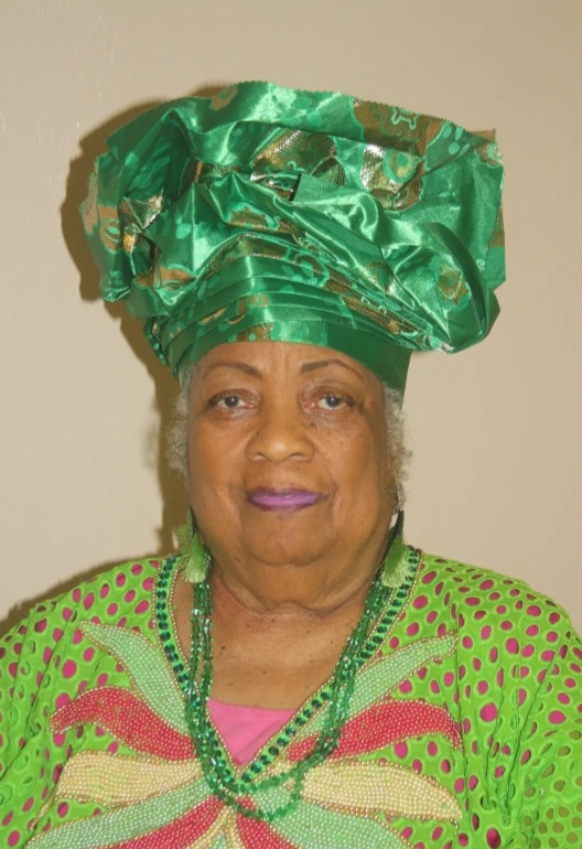 A person with grey hair wearing a shimmery green headdress, light green dangly earrings, and a green beaded necklace looks towards the camera. They wear a green and pink top with a pale green, pink, and yellow beaded embellishment.