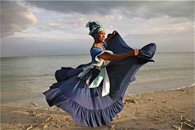 Marisol Blanco stands on a beach in a blue dress and headdress with the wind blowing her skirt